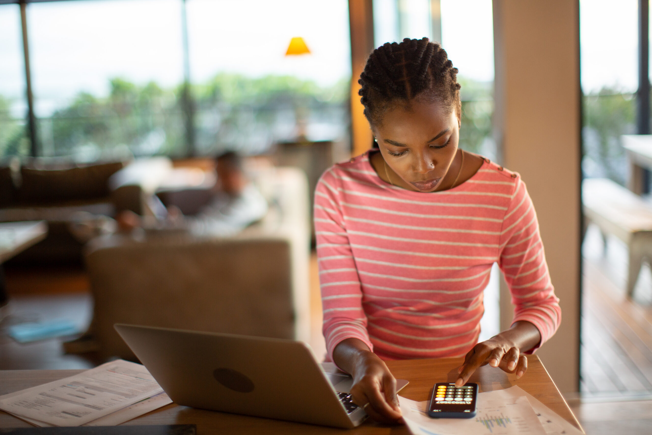 A woman with braided hair wearing a pink and white striped shirt sits at a wooden table, focused on a smartphone calculator. In front of her are a laptop and several printed financial documents. The background shows a dimly lit, modern living room with another person blurred in the distance.